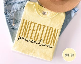 May include: A light yellow t-shirt with the words "INFECTION prevention" printed in gold and black. The shirt is folded and placed on top of blue jeans and a tan hat. A small circle with the word "BUTTER" is in the bottom right corner.