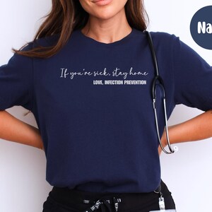 Puede incluir: Camiseta azul marino con el texto "If you're sick, stay home. Love, Infection Prevention" en escritura blanca. Un estetoscopio cuelga del cuello. La camiseta es de color s&oacute;lido.