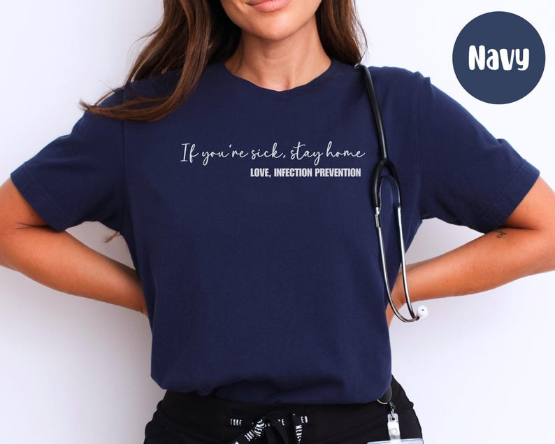 Puede incluir: Camiseta azul marino con el texto "If you're sick, stay home. Love, Infection Prevention" en escritura blanca. Un estetoscopio cuelga del cuello. La camiseta es de color s&oacute;lido.