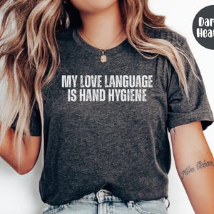 May include: Dark heather gray t-shirt with the text "MY LOVE LANGUAGE IS HAND HYGIENE" in white. The shirt has a crew neck and short sleeves. A dark gray circle with the words "Dark G. Heather" is in the upper right corner.