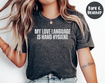 May include: Dark heather gray t-shirt with the text "MY LOVE LANGUAGE IS HAND HYGIENE" in white. The shirt has a crew neck and short sleeves. A dark gray circle with the words "Dark G. Heather" is in the upper right corner.