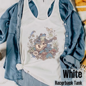 May include: White racerback tank top with a blue frog playing a guitar in a floral setting.