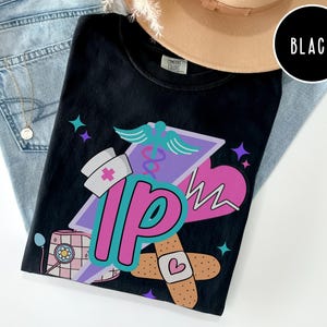 May include: Black t-shirt with a colorful graphic design featuring a nurse's hat, a medical symbol, a heart, and the letters "IP". The design includes a bandage with a heart and a stethoscope. The shirt is displayed on a pair of blue jeans.