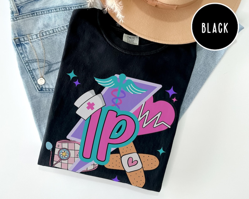 May include: Black t-shirt with a colorful graphic design featuring a nurse's hat, a medical symbol, a heart, and the letters "IP". The design includes a bandage with a heart and a stethoscope. The shirt is displayed on a pair of blue jeans.