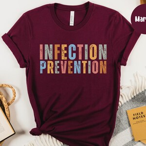 May include: Maroon t-shirt with the words "INFECTION PREVENTION" in colorful, distressed block letters. The shirt is displayed on a neutral background with a coffee cup, notebooks, and a woven basket.