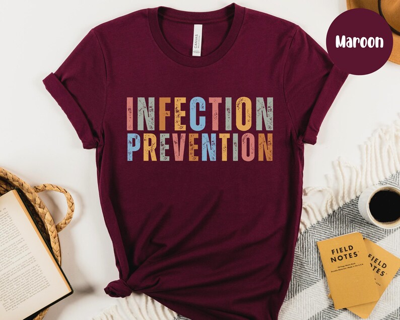 May include: Maroon t-shirt with the words "INFECTION PREVENTION" in colorful, distressed block letters. The shirt is displayed on a neutral background with a coffee cup, notebooks, and a woven basket.