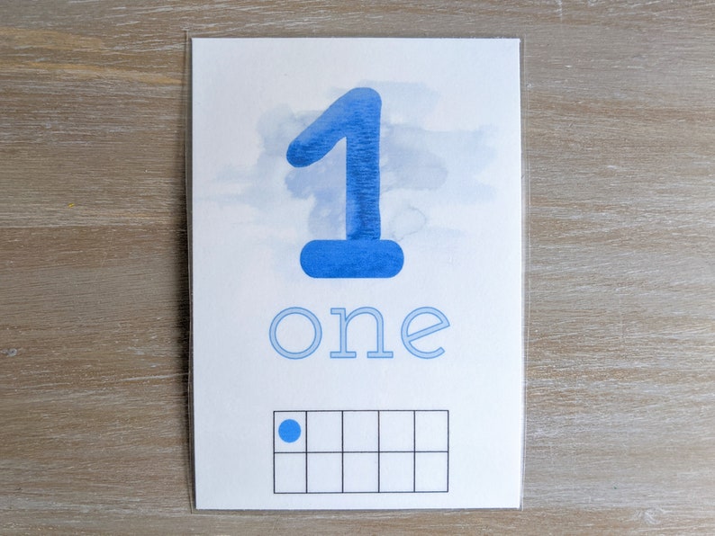 Pu&ograve; includere: Una carta rettangolare bianca con un grande numero 1 blu in stile acquerello in alto, la parola one scritta in lettere minuscole azzurre sotto e una griglia da dieci spazi in basso con un singolo punto blu nel primo quadrato.