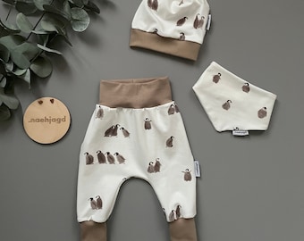 May include: A baby outfit set with a white and brown penguin print. The set includes a hat, pants, and a bib.