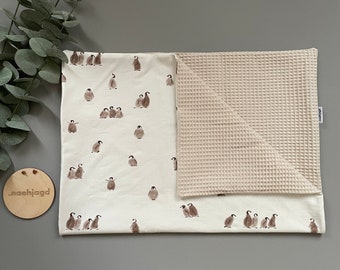 May include: A white baby blanket with a penguin pattern on one side and a waffle texture on the other side. The blanket is folded in half and has a small tag with the word "maehijagd" on it.