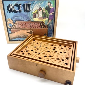 May include: A wooden Space Tilt labyrinth board game featuring a tan maze surface with numbered holes and a metal ball. The game includes a cardboard box with the title Space Tilt and an illustration of people playing the game. The board has two wooden knobs on the sides for tilting the maze to navigate the ball through the path.
