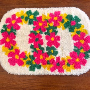May include: A rectangular tufted rug with a cream-colored background featuring a repeating pattern of bright pink, yellow, and green flowers. The floral design is arranged in two circular clusters, creating a soft, textured surface with a high pile.
