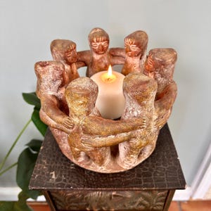 May include: A circular terracotta candle holder featuring six stylised figures standing in a ring with their arms linked around a central white pillar candle. The figures have a textured, earthy brown finish with visible highlights and shadows.