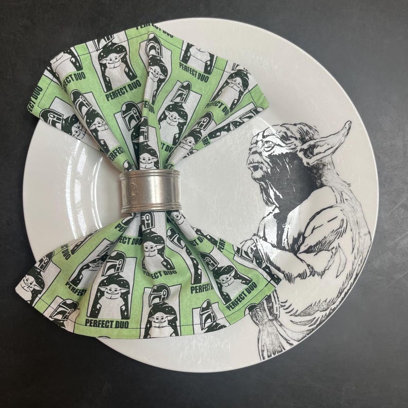 Puede incluir: Un plato de cer&aacute;mica blanco con un gr&aacute;fico de Yoda en blanco y negro. Una servilleta estampada en verde y blanco con el texto "PERFECT DUO" se sujeta con un servilletero plateado. La servilleta presenta im&aacute;genes de Baby Yoda y un casco mandaloriano.
