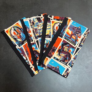Puede incluir: Fundas para cuadernos con un collage de im&aacute;genes de la serie The Mandalorian. Las fundas tienen un borde negro e ilustraciones de personajes en azul, naranja y beige.