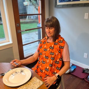 May include: An orange bib featuring a repeating pattern of black cat faces and spiderwebs. The bib is worn by a person seated at a wooden table, with a plate and napkin visible. The bib is likely for Halloween or a similar themed event.
