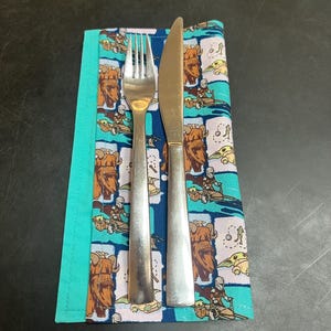 Puede incluir: Un juego de tenedor y cuchillo de acero inoxidable sobre una servilleta de tela con tem&aacute;tica de Star Wars. La servilleta presenta un patr&oacute;n repetido de personajes en tonos marrones, azules y verdes. La servilleta tiene un borde turquesa.