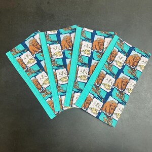 Puede incluir: Juego de cinco posavasos de tela con un patr&oacute;n repetido de una criatura marr&oacute;n sobre un fondo azul y turquesa. Cada posavasos tiene un borde turquesa. Los posavasos est&aacute;n dispuestos en forma de abanico.