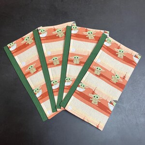Puede incluir: Cuatro cuadernos de tela con un patr&oacute;n repetido de un personaje alien&iacute;gena de piel verde sobre un fondo a rayas crema y naranja. Los cuadernos tienen lomo verde.