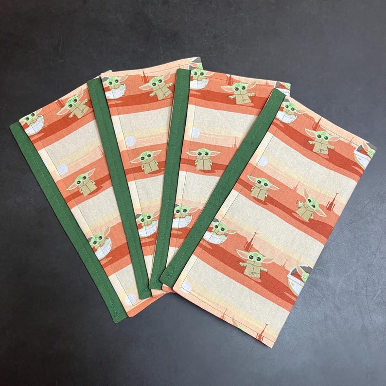 Puede incluir: Cuatro cuadernos de tela con un patr&oacute;n repetido de un personaje alien&iacute;gena de piel verde sobre un fondo a rayas crema y naranja. Los cuadernos tienen lomo verde.