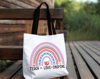 May include: A white tote bag with black handles featuring a colorful rainbow graphic composed of pencils and hearts. A red apple icon sits at the center of the rainbow. The text "TEACH LOVE INSPIRE" is printed in black below the graphic.