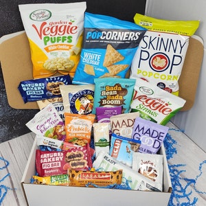May include: A white gift box filled with a variety of snack foods, including popcorn, veggie puffs, granola bars, and fig bars. The box is surrounded by blue tissue paper.