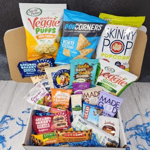 May include: A white box filled with a variety of snacks, including veggie puffs, popcorn, granola, and fig bars. The box is surrounded by blue tissue paper.