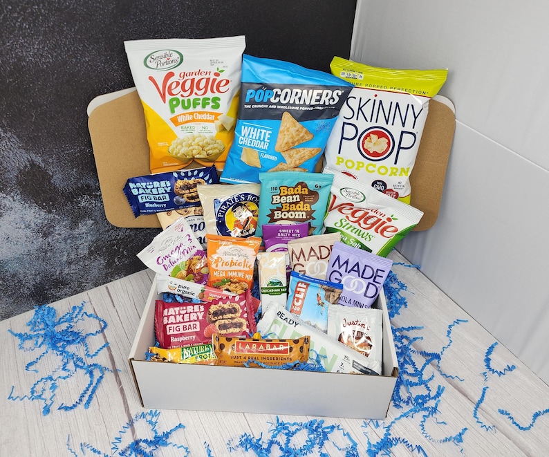 May include: A white gift box filled with a variety of snack foods, including popcorn, veggie puffs, granola bars, and fig bars. The box is surrounded by blue tissue paper.