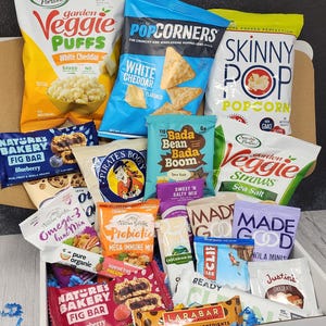 May include: A white box filled with a variety of snacks, including veggie puffs, popcorn, granola bars, fig bars, and other healthy snacks. The box is filled to the brim with snacks.