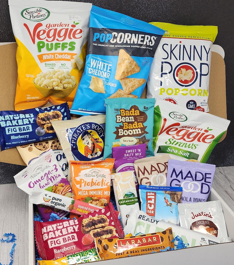 May include: A white box filled with a variety of snacks, including veggie puffs, popcorn, granola bars, fig bars, and other healthy snacks. The box is filled to the brim with snacks.