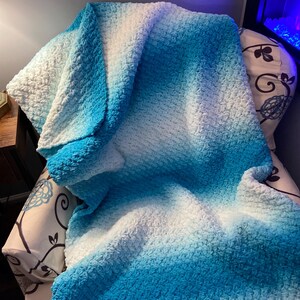 May include: A soft, crocheted blanket in shades of white and blue, transitioning from light to a deep turquoise. The blanket is draped over a chair, showcasing its textured surface and gradient colour scheme. The cosy, handmade blanket is perfect for home decor.