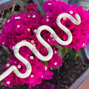 May include: A cream-colored, serpentine-shaped decorative object with light blue and gold accents, resting on a bed of vibrant magenta bougainvillea flowers. The flowers have small white centers, creating a striking contrast.