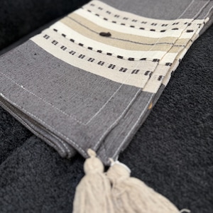 Peut inclure: Textile plié gris et crème avec un motif à rayures et des glands. Le design comprend des rayures horizontales grises, crème et beige, accentuées de petits tirets noirs. Deux glands crème pendent du bord.