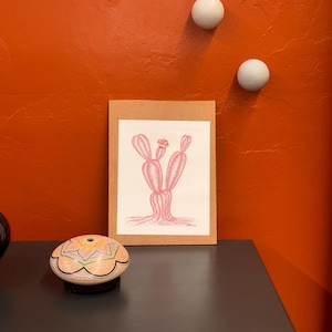 Puede incluir: Un dibujo de cactus rosa en un marco marrón está sobre una superficie oscura. Una pieza decorativa de cerámica con patrones coloridos está en primer plano. El fondo presenta una pared naranja con dos decoraciones murales esféricas blancas.
