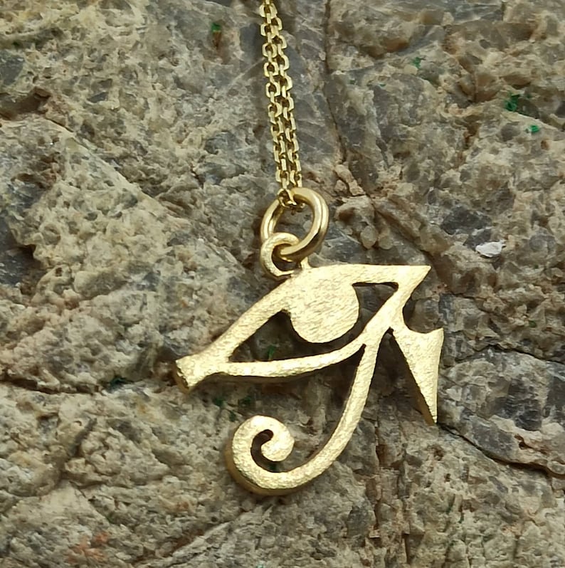 May include: A gold-colored pendant necklace featuring an Egyptian Eye of Horus symbol. The pendant is detailed and intricate, with a swirl design at the bottom.