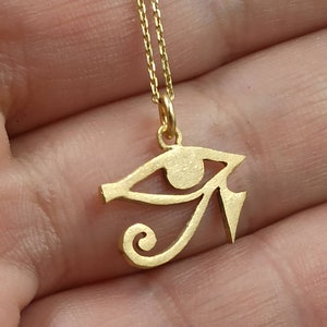 May include: A gold-colored necklace with a pendant shaped like the Eye of Horus, an ancient Egyptian symbol of protection.
