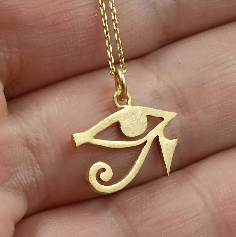 May include: A gold-colored necklace with a pendant shaped like the Eye of Horus, an ancient Egyptian symbol of protection.