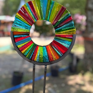 Könnte beinhalten: Ein farbenfrohes, kreisförmiges Glaskunstwerk mit einem Regenbogen aus Farben, darunter Rot, Orange, Gelb, Grün und Blau. Das Glas ist in einem Metallrahmen gefasst und auf zwei Metallstäben montiert.