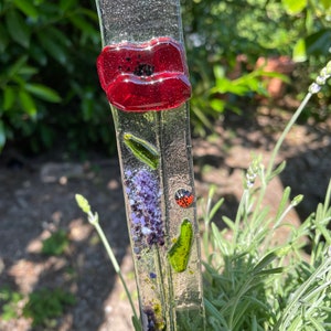 Può includere: Un paletto da giardino in vetro con un fiore di papavero rosso, foglie verdi, fiori di lavanda viola e una coccinella. Il paletto è in vetro trasparente con una superficie testurizzata.