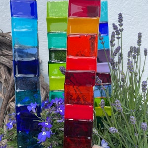 Op de afbeelding: Een set van vier kleurrijke glazen tuinstokken, elk gemaakt van gestapelde glazen tegels in verschillende kleuren. De stokken zijn in een rij gerangschikt, met de hoogste stok in het midden. De stokken zijn omringd door groene planten en paarse bloemen.