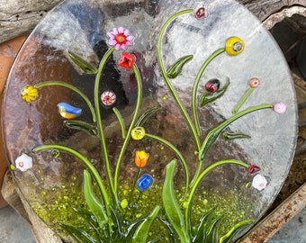 May include: A round glass suncatcher with a floral design. The suncatcher features green leaves, colourful flowers, and small glass beads. It is hanging from a string.