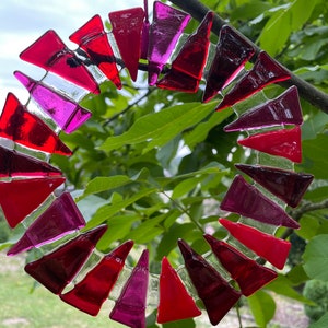 Może przedstawiać: Okrągły witrażowy łapacz słońca z różnymi czerwonymi i różowymi trójkątnymi elementami. Szkło ułożone jest w spiralny wzór, zawieszone na cienkim, różowym sznurku. Tło to rozmycie zielonych liści.