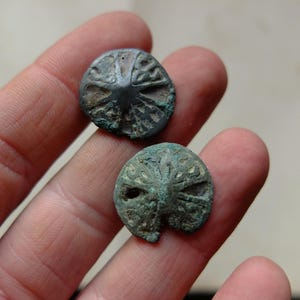 Puede incluir: Dos botones metálicos circulares antiguos con intrincados diseños en relieve. Los botones son de un color oscuro y envejecido con toques de pátina verde, lo que sugiere antigüedad. Los botones se sostienen en una mano.