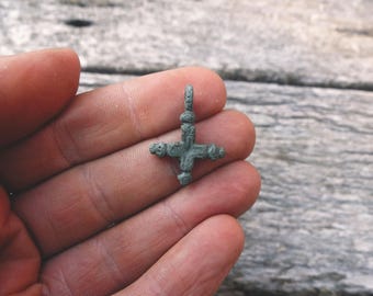 May include: A small, weathered, green-toned metal cross pendant featuring a textured surface, a central vertical bar with a loop at the top, and three rounded, decorative ends on the horizontal and bottom arms of the cross.