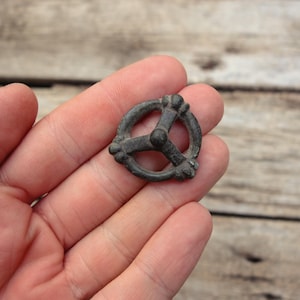 Puede incluir: Pequeño objeto metálico antiguo, de color gris oscuro, con un diseño circular y tres brazos. El objeto tiene un aspecto desgastado, lo que sugiere antigüedad. El diseño incluye elementos decorativos en los extremos de los brazos y alrededor del círculo. El objeto se sostiene en una mano.