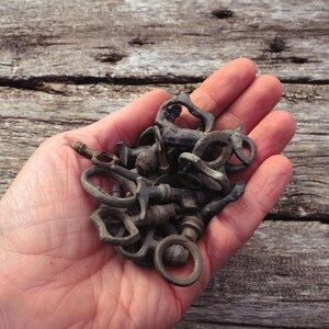 May include: A collection of antique bronze keys and key components, in various shapes and sizes, are held in an open hand. The keys have a weathered, aged appearance, suggesting they are vintage or reclaimed. The background is a rustic wooden surface.