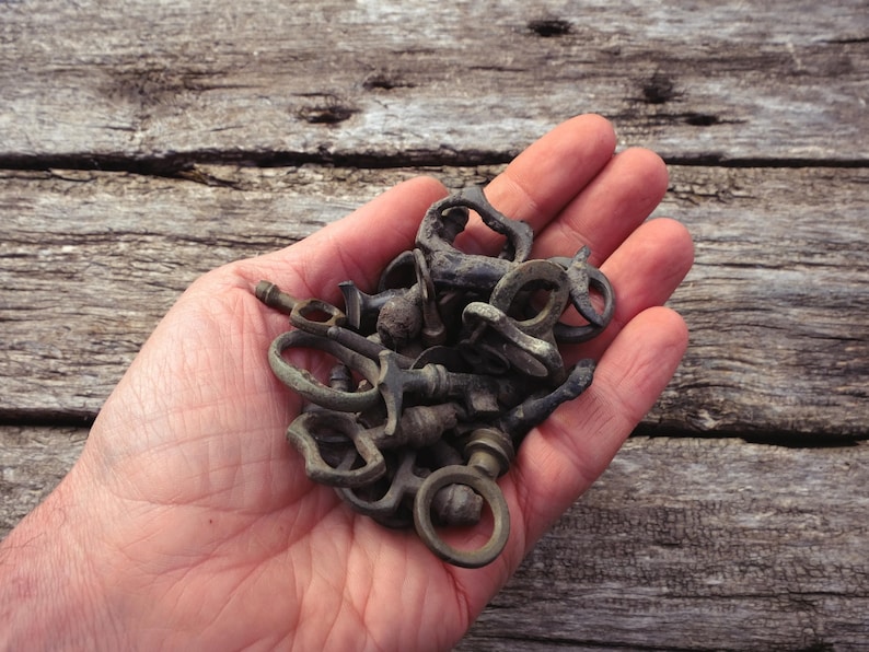 May include: A collection of antique bronze keys and key components, in various shapes and sizes, are held in an open hand. The keys have a weathered, aged appearance, suggesting they are vintage or reclaimed. The background is a rustic wooden surface.