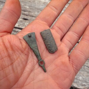 May include: Two small, weathered bronze-colored metal artifacts. One piece is a triangular-shaped tool with a circular loop at the narrow end and a small stud at the top. The second piece is a rectangular, textured metal fragment with worn edges.
