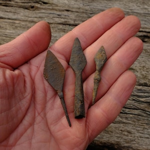 Könnte beinhalten: Drei antike, rostige Metallpfeilspitzen in verschiedenen Formen und Größen werden in einer offenen Hand gezeigt. Die Pfeilspitzen haben ein verwittertes Aussehen, was auf Alter und historische Bedeutung hindeutet. Der Hintergrund ist eine verwitterte Holzoberfläche.