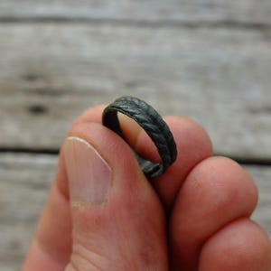 以下が含まれることがあります： 編み込み模様のダークグレーのリング。指の間にリングが挟まれ、その質感と細部が際立っています。リングの表面は風化しているように見え、アンティークまたはヴィンテージスタイルを示唆しています。リングはジュエリーです。