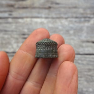May include: A small, dark metallic thimble featuring a textured, dimpled surface across the dome and a smooth, banded base. The object has a weathered, aged appearance with a muted, brownish-grey finish and a cylindrical shape with a rounded top.